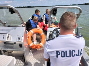Zdjęcie przedstawia: umundurowanego policjanta za sterem łodzi, z tyłu widoczne dzieci w kapokach.