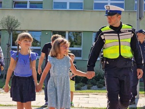 Zdjęcie przedstawia dwie dziewczynki i policjant idą trzymając się za ręce.