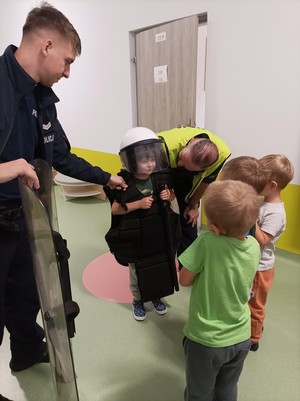 Zdjęcie przedstawia: dwóch umundurowanych policjantów podczas prezentacji policyjnego sprzętu. Dzieci stoją wokół mundurowych i oglądają kamizelkę i kask policyjny.
